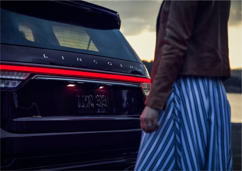 A person is shown near the rear of a 2023 Lincoln Aviator® SUV as the Lincoln Embrace illuminates the rear lights | Mark McLarty Lincoln in North Little Rock AR
