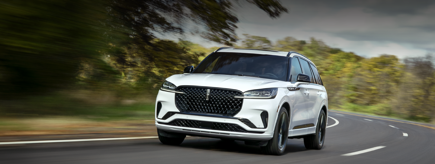 A white 2025 Lincoln Aviator® SUV is being driven down a road lined with trees. | Mark McLarty Lincoln in North Little Rock AR